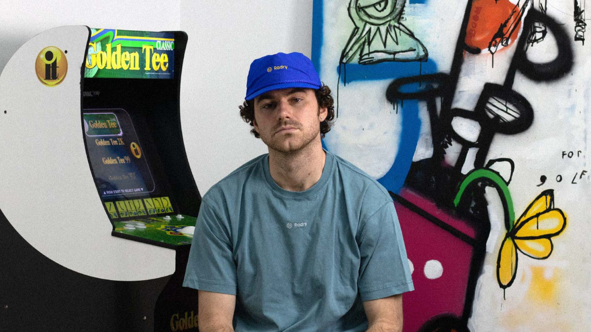 Man in a blue Radry cap and teal T-shirt sitting by a graffiti wall and a Golden Tee arcade machine
