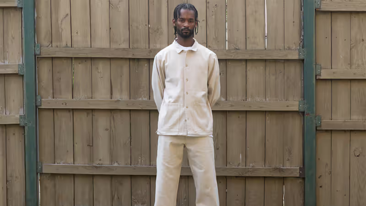 Man in a cream Whim Golf jacket and matching trousers standing in front of a wooden fence