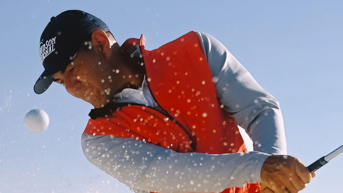 Golfer in an orange vest and navy cap splashing a ball out of a bunker
