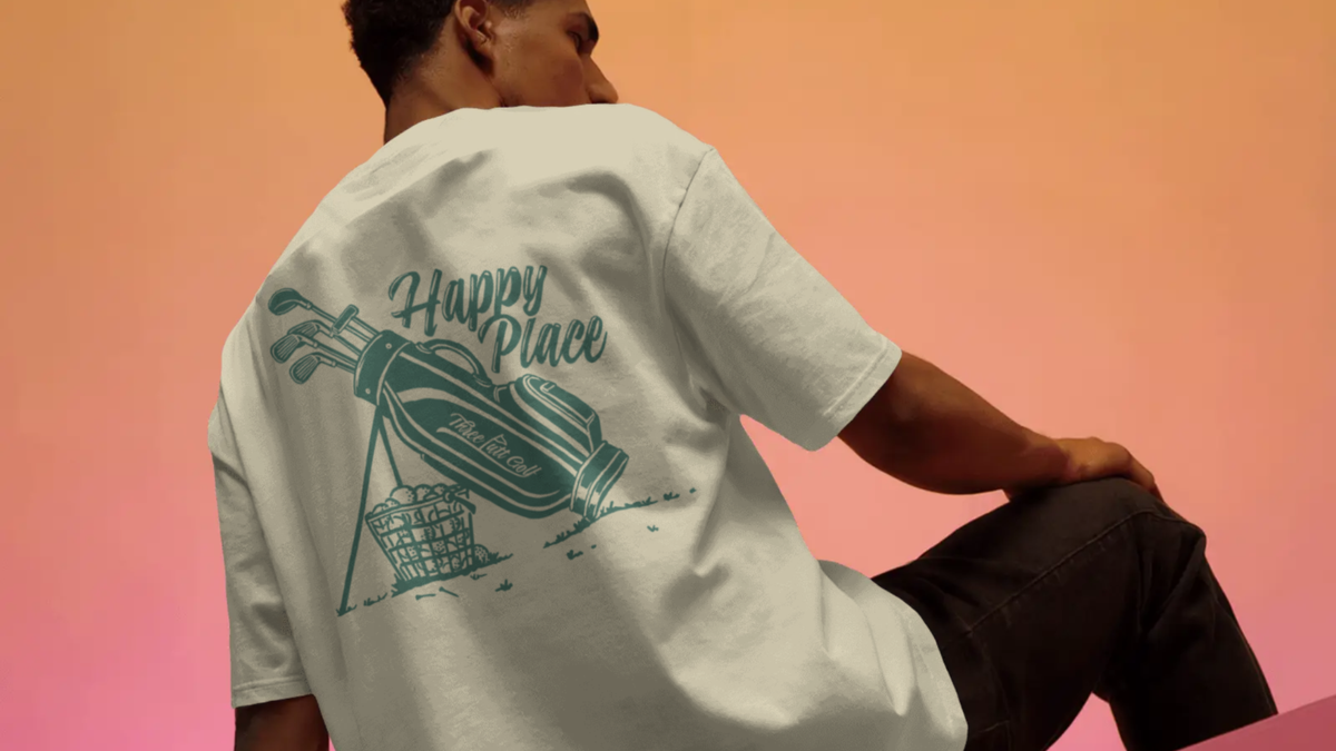 Man seated, seen from behind in a Happy Place golf T-shirt in Cream, showing the golf bag and Happy Place back print