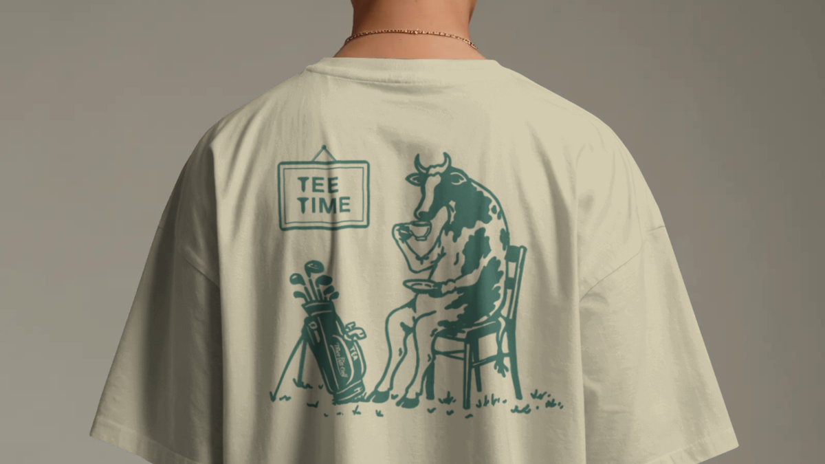 Man seen from behind in a Tee Time golf T-shirt in Cream, showing the cow drinking tea under a Tee Time sign
