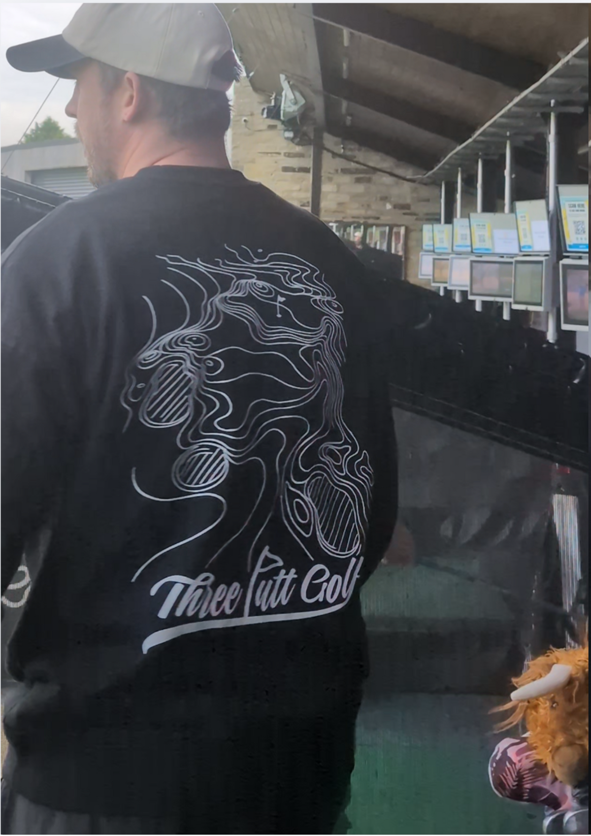 Golfer at the driving range in the Contours golf sweatshirt in Black from behind, showing the topographic map print and Three Putt Golf script