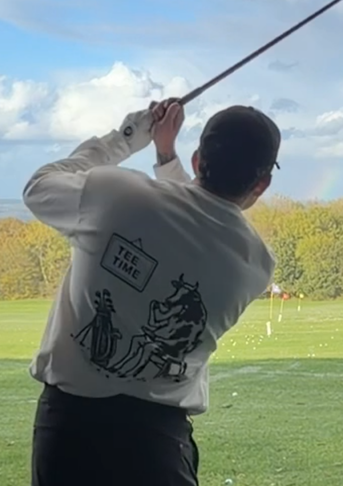 Close-up of the Tee Time golf long-sleeve tee mid-swing at the driving range, showing the Tee Time sign, golf bag and cow drinking tea on the back