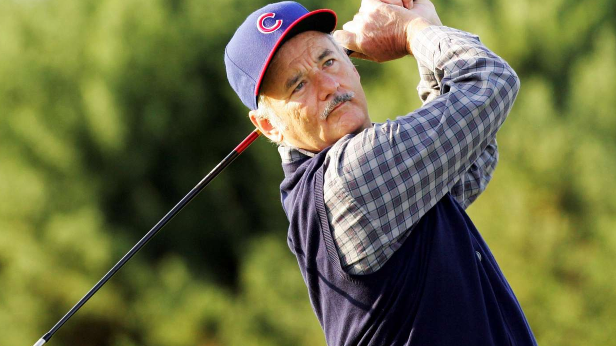 Bill Murray swinging a golf club in a Chicago Cubs cap, plaid shirt and navy sweater vest