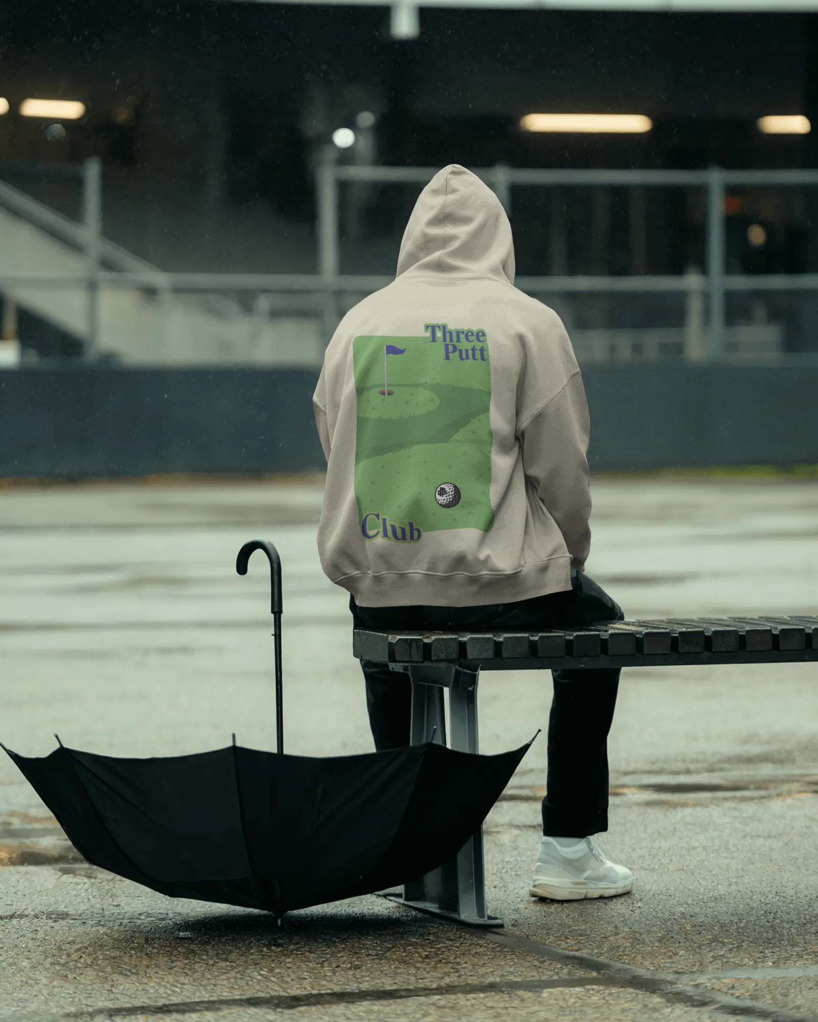 Person wearing a hoodie with a golf-themed design, sitting on a bench with an umbrella in a parking lot.
