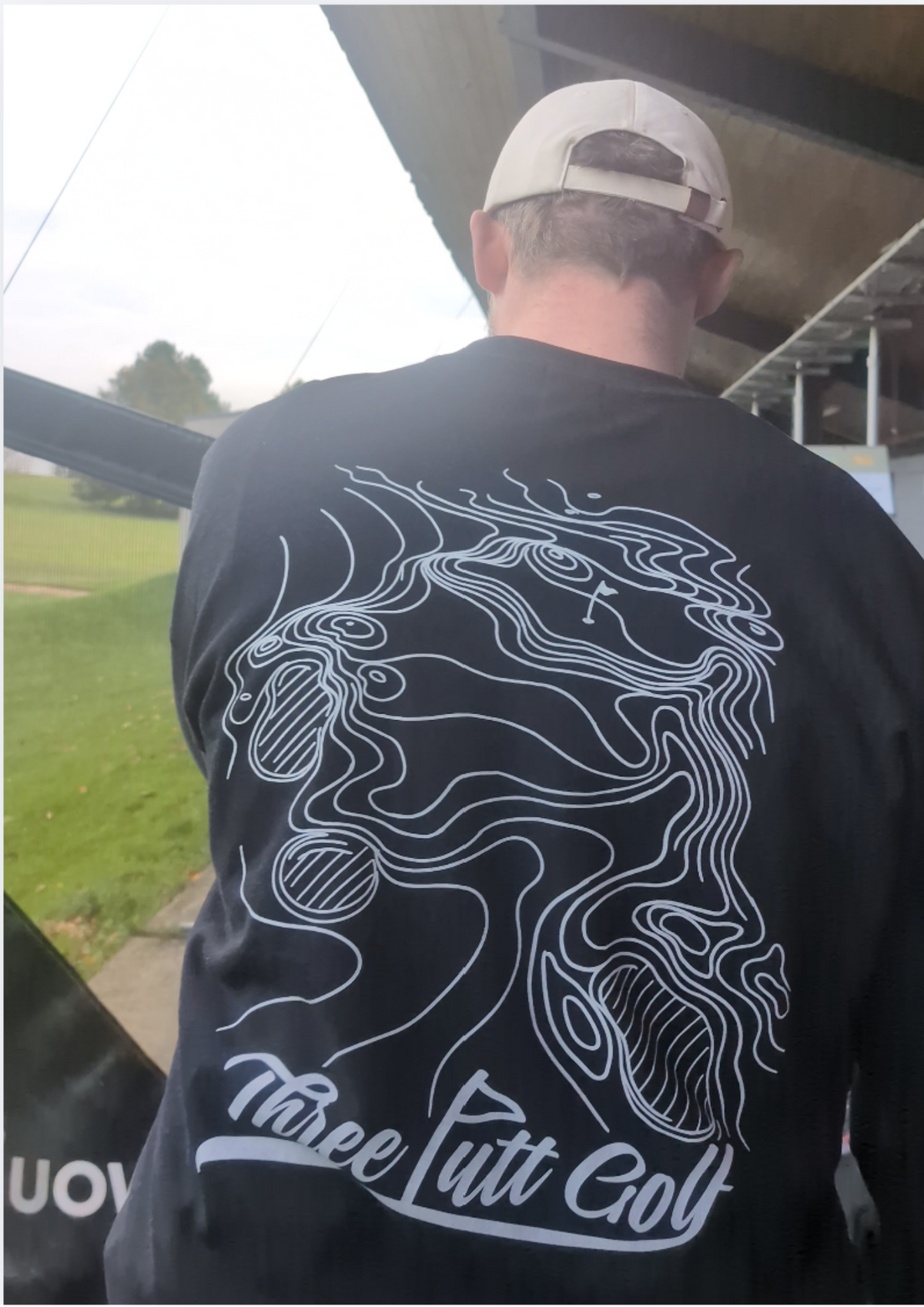Golfer at the driving range in the Contours golf sweatshirt in Black seen from behind, showing the full topographic map print and Three Putt Golf script