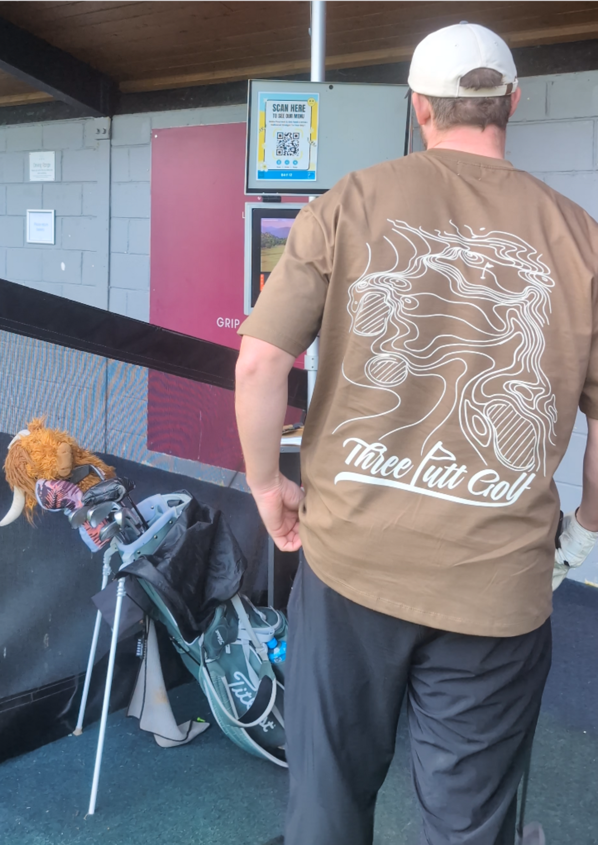 Golfer in the Contours golf T-shirt in Brown seen from behind beside a golf bag at the driving range, showing the topographic map print