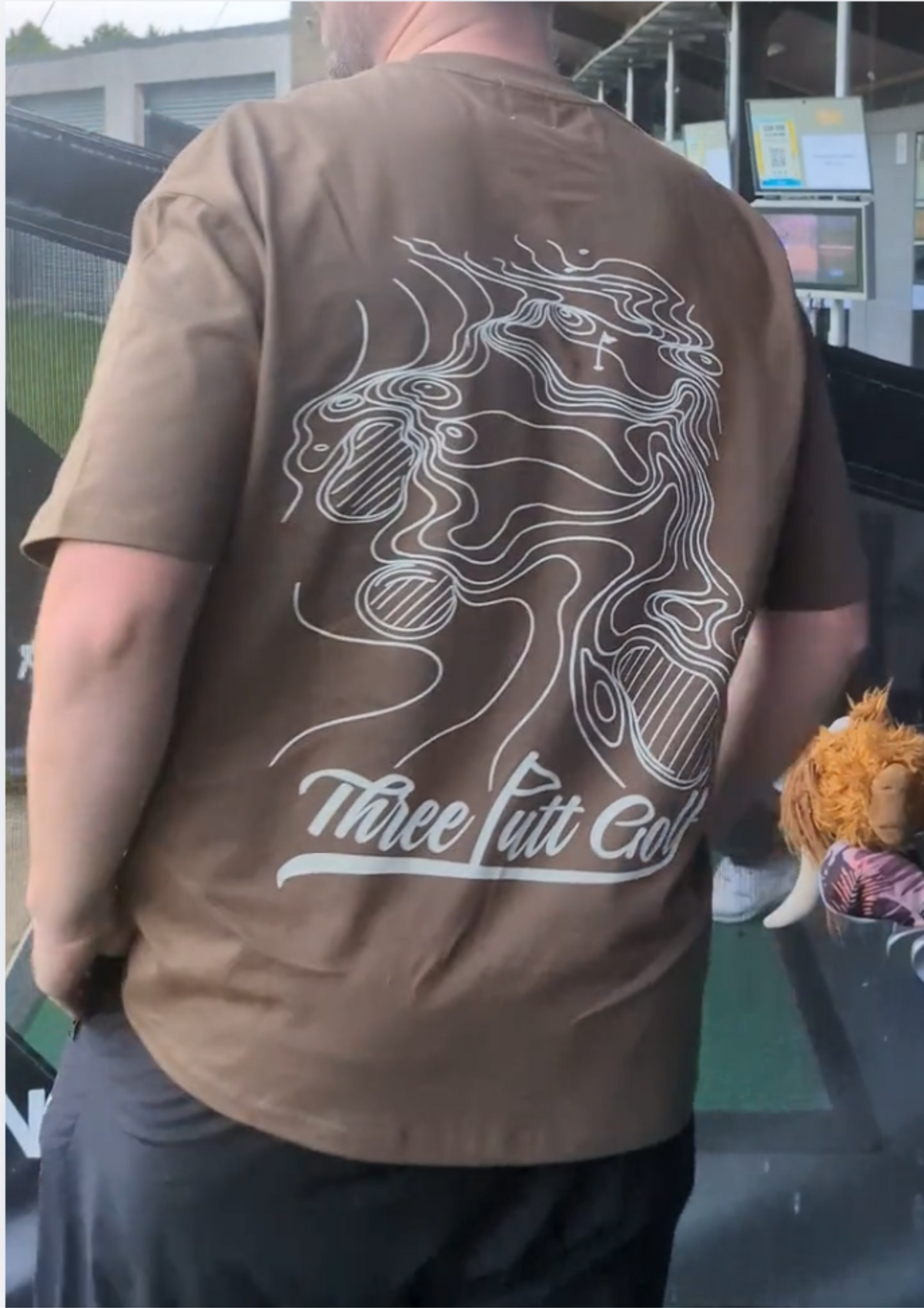 Back of the Contours golf T-shirt in Brown worn at the driving range, with the topographic green print and Three Putt Golf script
