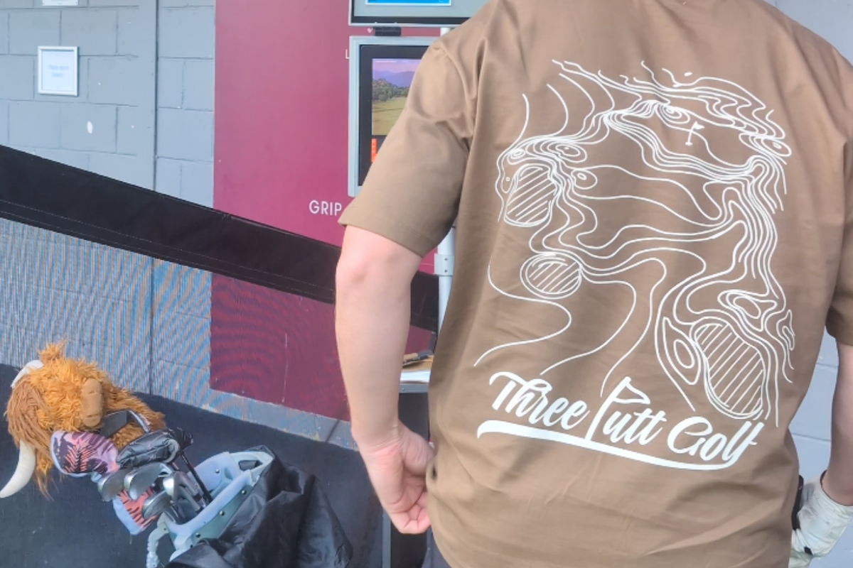 Golfer in the Contours golf T-shirt in Brown seen from behind beside a golf bag at the driving range, showing the topographic map print