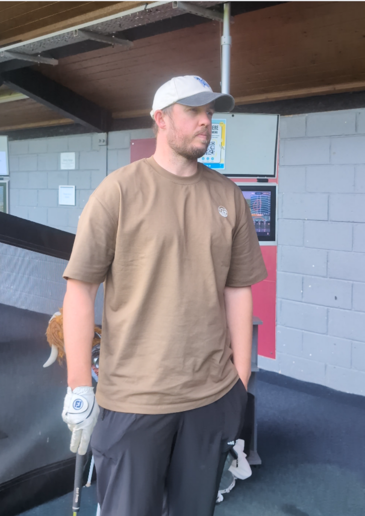 Golfer in a driving range bay wearing the Contours golf T-shirt in Brown, front view with the round cow badge on the chest