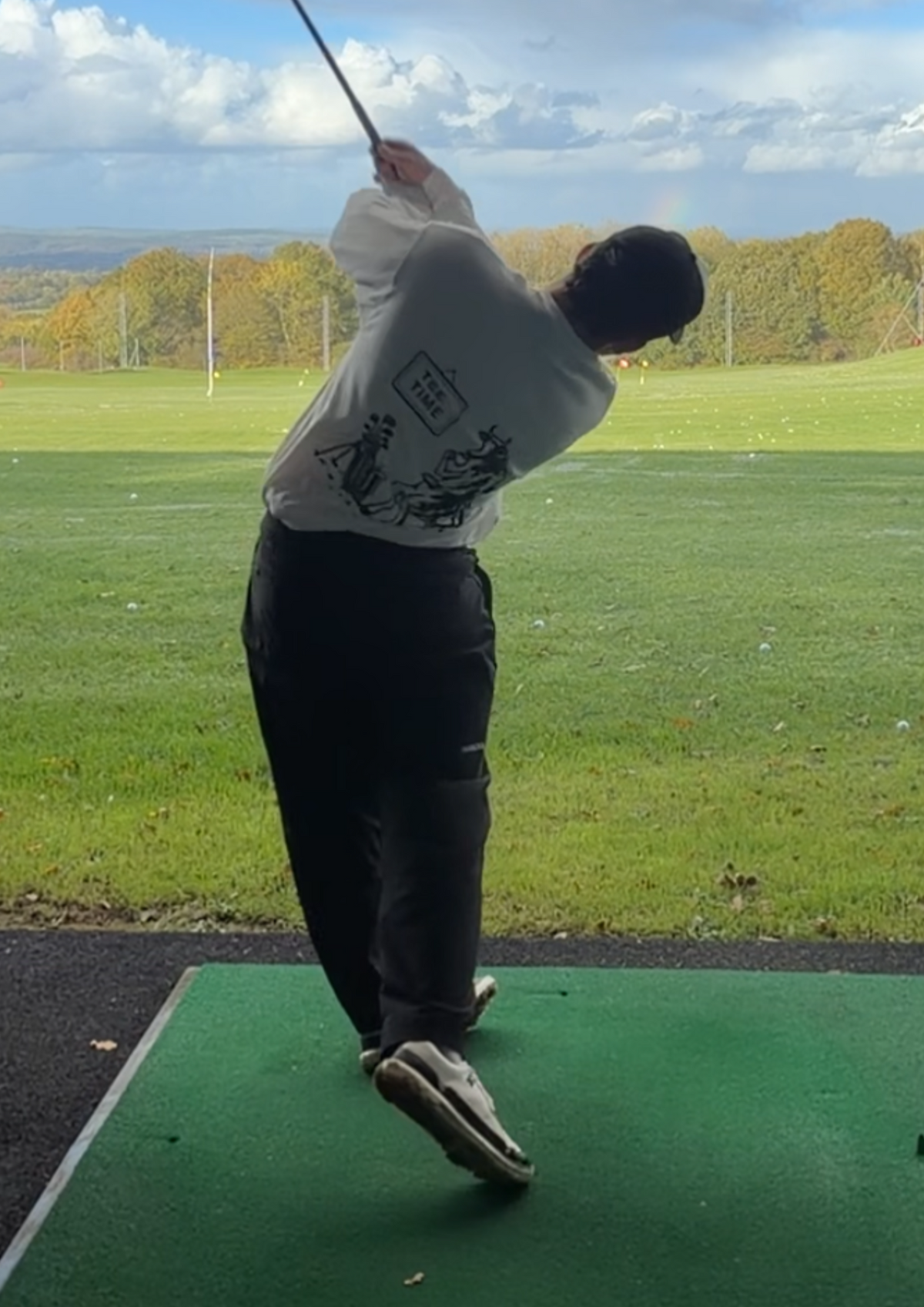 Golfer mid-swing at the driving range in the Tee Time golf long-sleeve tee, showing the cow having a cup of tea beside its clubs on the back